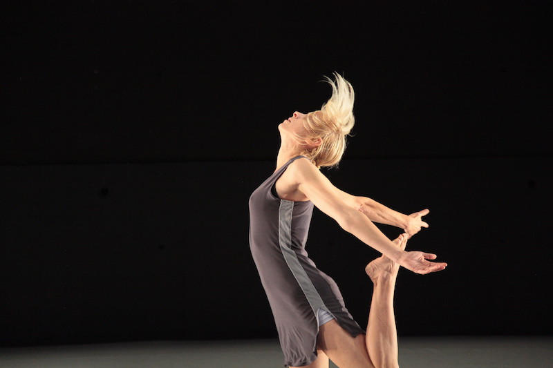 Liz Gerring in a racer back athletic-looking tunic kicks her leg behind her. Her head looks to the ceiling as her short blonde hair tosses up.